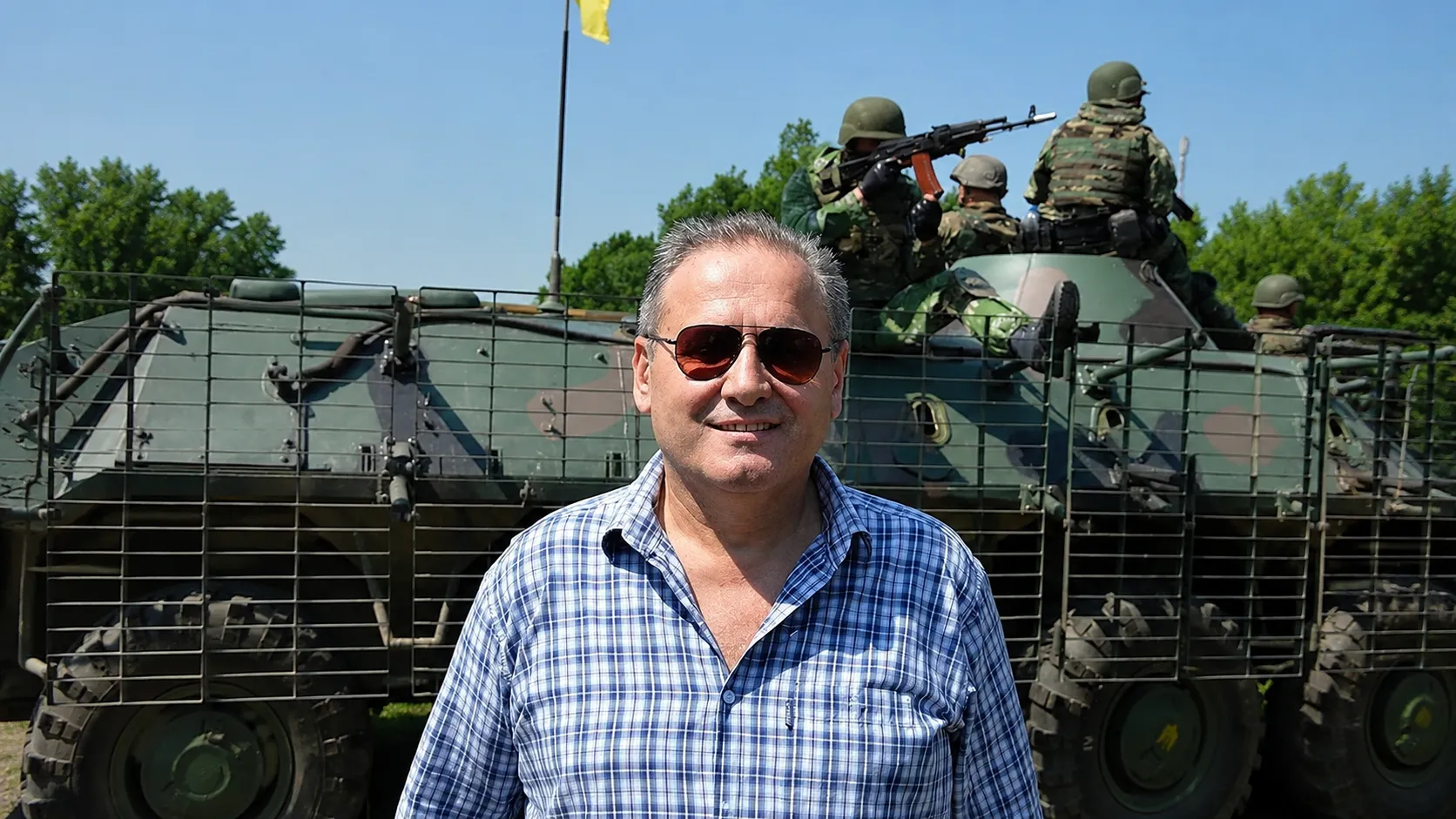 Богдан Кушнір у сонцезахисних окулярах на тлі бронетранспортера, на броні — військові зі зброєю, поряд майорить український прапор